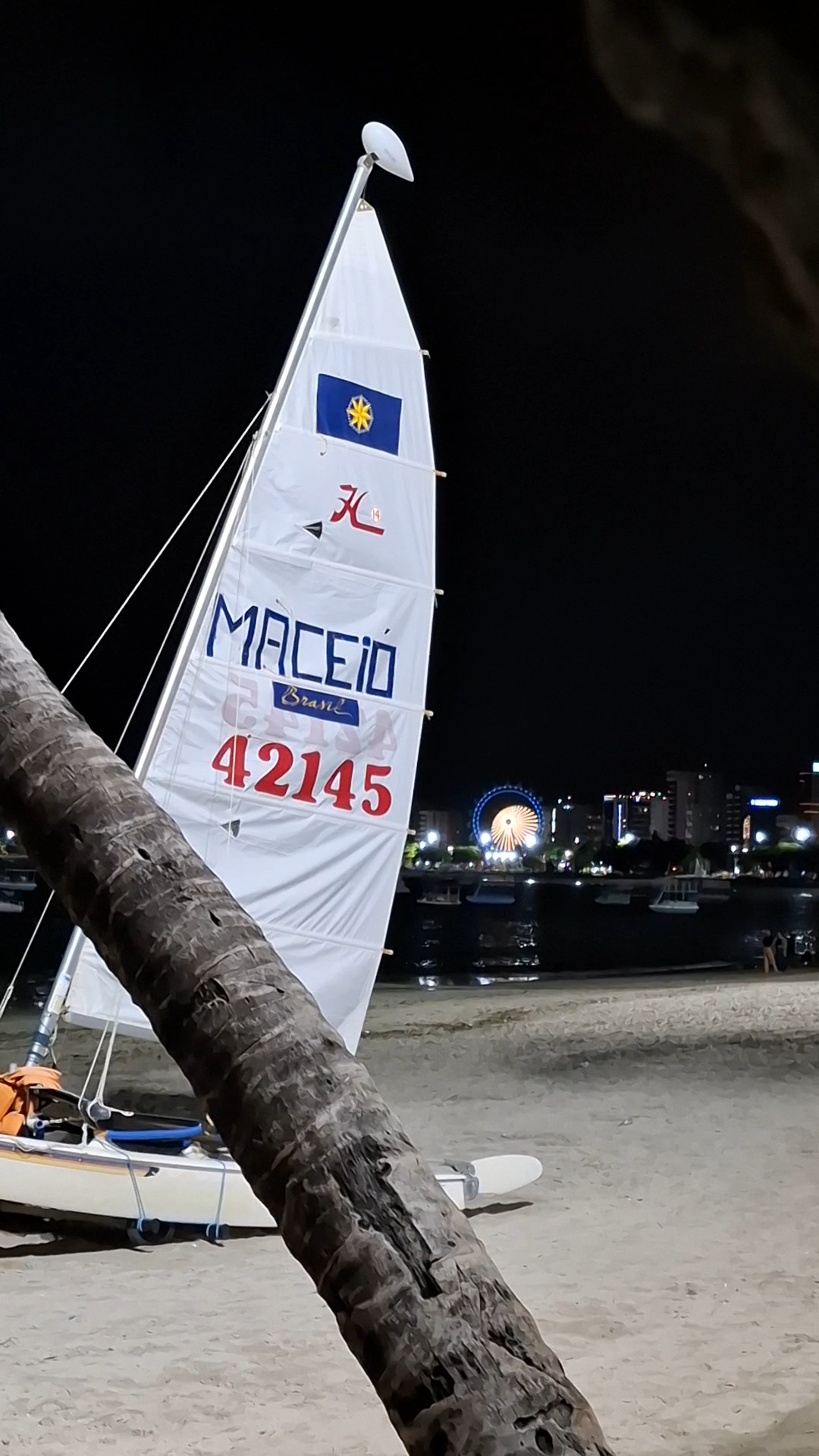 Playa de Maceió en la noche, vela desplegada del bote que dice Maceió. Rueda gigante al fondo.